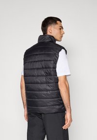 Black, sleeveless puffer vest with a high collar, featuring quilted padding and a smooth texture, worn over a white t-shirt.