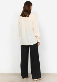 Cream blouse with smocked detailing at the shoulders, paired with loose black trousers. Smooth fabric and relaxed silhouette.