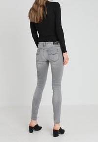 Mujer vestida con jeans ajustados gris claro, top negro de manga larga y sandalias negras de tacón cuadrado, de pie contra un fondo claro y liso.