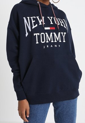 Donkerblauwe hoodie met de tekst en het logo "New York Tommy Jeans", gedragen met een blauwe spijkerbroek, met trekkoorden en een voorkantzak.