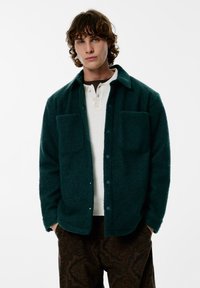 Giacca in pile di colore verde petrolio scuro con texture, con due tasche anteriori e bottoni blu, indossata sopra una polo bianca e pantaloni marroni con motivo.