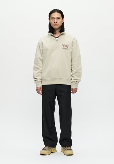 Uomo con lunghi capelli scuri che indossa una felpa beige con zip a metà, pantaloni neri e scarpe da trekking beige, in piedi su uno sfondo bianco.