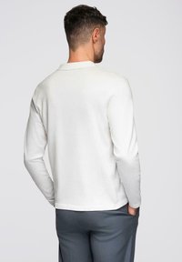 Witte langlopende blouse, gemaakt van een zachte stof, met een ronde halslijn en een rechte zoom. De achterkant toont een minimalistisch ontwerp zonder versieringen.