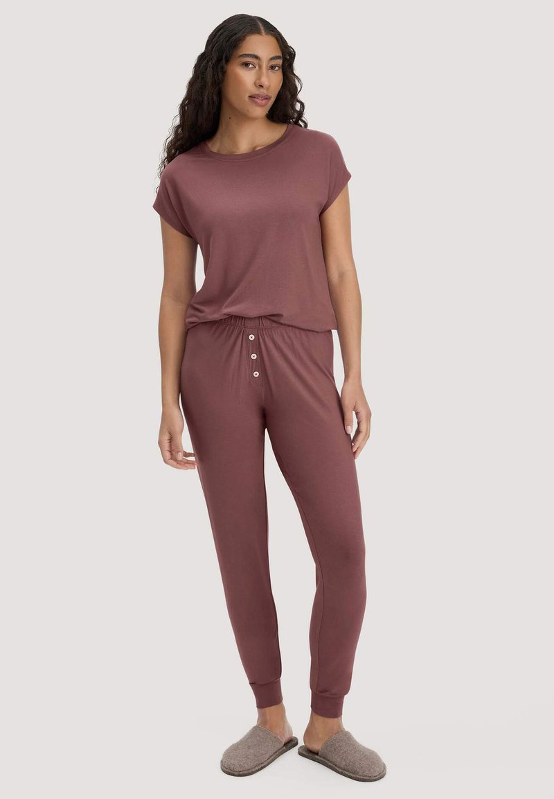 Donna che indossa una maglietta a maniche corte color malva e pantaloni jogger coordinati con bottoni, in piedi a piedi nudi con pantofole grigie senza lacci.
