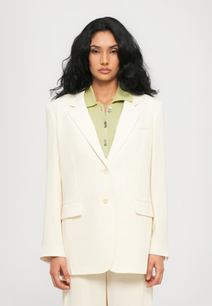 Crème-kleurige blazer met een gestructureerde vorm, voorzien van een dubbele knoopsluiting, zijzakken en een gladde textiele afwerking. Daaronder een groene geribde polo.