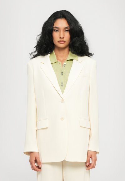 Cremfarbener Blazer mit strukturierter Form, ausgestattet mit einem Zwei-Knopf-Verschluss, Seitentaschen und einem glatten Textilfinish. Darunter ein grünes geripptes Poloshirt.