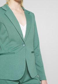 Zelené sakové sako vyrobené z hladkého materiálu. Obsahuje jeden knoflík, klopové límce a kontrastní černé kapsy. Úzký střih.