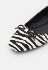 Płaskie buty w zebra wzór w czerni i bieli, z miękką futrzaną teksturą, zaokrąglonym czubkiem oraz małym czarnym kokardką z srebrnym akcentem.