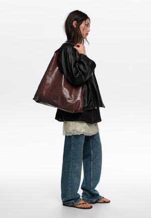 Mujer con chaqueta de cuero negra, top crema con encaje, vaqueros holgados, sandalias, llevando un gran bolso tote de cuero marrón oscuro al hombro.