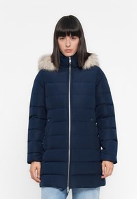 Giubbotto piumino blu navy con cappuccio foderato in pelliccia, dotato di zip frontale, design trapuntato, due tasche laterali e un taglio di lunghezza media.