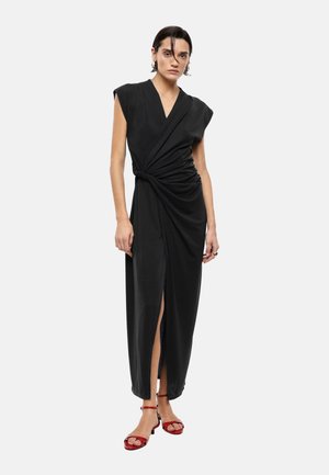 Donna che indossa un abito nero avvolgente senza maniche con spacco laterale e tacchi rossi aperti sulle dita, in piedi davanti a uno sfondo chiaro e semplice.