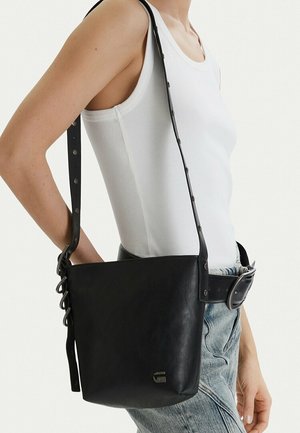 Persona vistiendo una camiseta sin mangas blanca y jeans azules llevando un bolso de cuero negro con una correa ajustable.