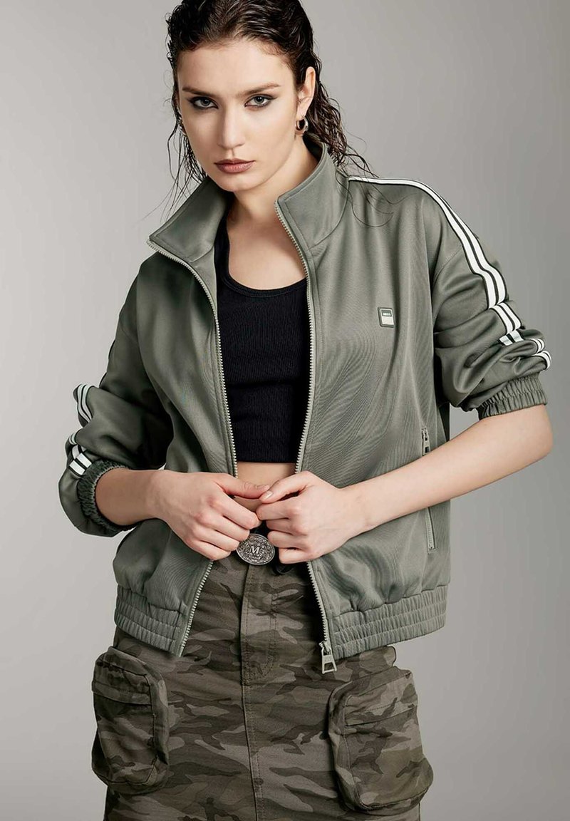 Chaqueta verde oliva con cremallera y rayas blancas en las mangas, puños de canalé y bolsillos laterales, combinada con una camiseta corta negra y una falda de camuflaje.