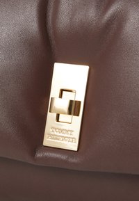 Ruda odinė rankinė su stačiakampiu auksinės spalvos užsegimu, ant kurio išgraviruota "Tommy Hilfiger" logotipas. Minkšta tekstūra ir struktūruota forma.