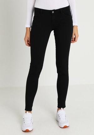 Jean slim noir porté avec des baskets blanches et un haut blanc à manches longues, visible de la taille aux pieds sur un fond clair uni.