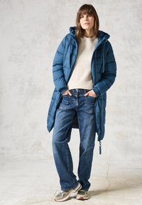 Vadderad teal-jacka med huva, krämfärgad texturerad tröja, blå jeans och grå sneakers. Avslappnad passform med en lång design och dragkedjedetaljer.
