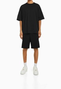 Osoba oblečená v volném černém tričku s krátkým rukávem, černých šortkách ke kolenům, bílých ponožkách a bílých teniskách stojí na bílém pozadí.