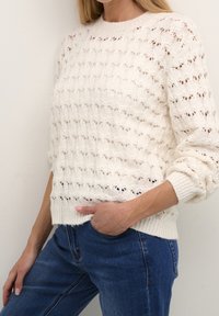 Pull en tricot blanc avec un motif ajouré détaillé, col rond et poignets côtelés. Associé à un jean bleu foncé.