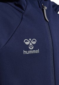 Marinblå sportjacka i stretchigt tyg med dragkedja framtill och en ljusgrå logotyp med abstrakt design på bröstet.