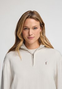 Cremfarbener Fleece-Pullover mit Viertelreißverschluss und weicher Textur, der ein kleines gesticktes Logo auf der Brust und einen Reißverschlussverschluss hat.