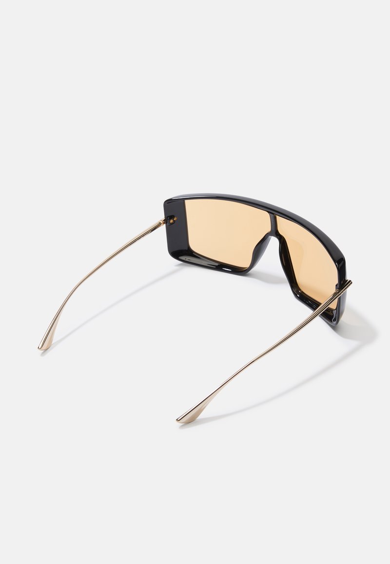 Sonnenbrille mit einem schwarzen Kunststoffrahmen, goldenen Metallbügeln und bernsteinfarbenen Gläsern. Das Design zeichnet sich durch eine geometrische Form und einen minimalistischen Stil aus.