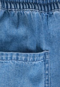 Κοντινή λήψη μπλε τζιν υφάσματος με ελαστική μέση και τετράγωνη μπροστινή τσέπη με ορατά ραμμένα ράμματα.