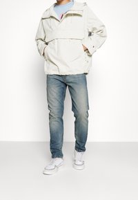 Giacca con cappuccio di un beige chiaro, dotata di cordini regolabili, tasca frontale e orlo elastico. Indossata con jeans blu chiaro e sneakers bianche.