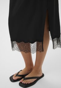 Jupe midi noire avec ourlet en dentelle florale et fente latérale, portée avec des sandales plates noires à brides tongs pieds nus.