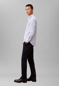 Chemise bleu clair à manches longues avec col et boutons, associée à un pantalon slim foncé et des chaussures noires, vue de profil.