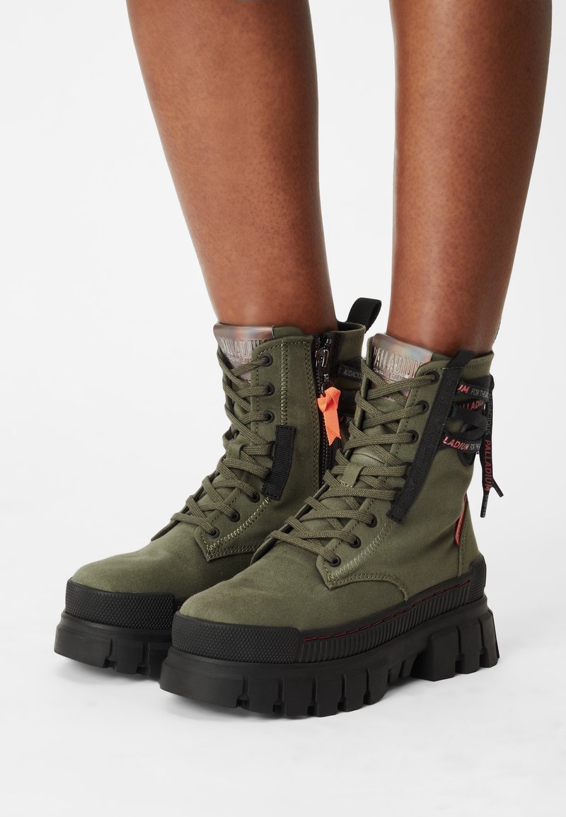 Bottes à lacets vert olive avec semelles épaisses noires, fermeture éclair latérale, et languettes de tirage noires et rouges portées sur jambes nues.