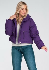 Skórzana fioletowa kurtka puchowa z kapturem, z dwoma zapinanymi na zamek kieszeniami z przodu i błyszczącym wykończeniem. Noszona na jasnoszarym swetrze.
