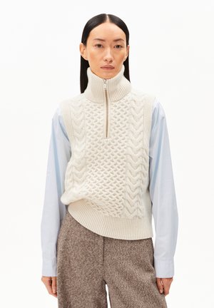 Pull sans manches en tricot torsadé couleur crème avec un col montant et une fermeture éclair haute. Porté par-dessus une chemise bleu clair, associé à un pantalon marron texturé.