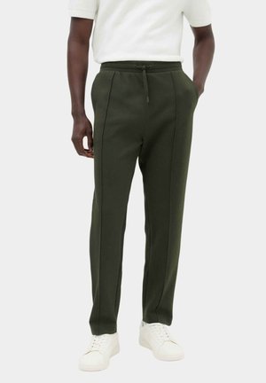 Hombre con pantalones verde oscuro con cordón, zapatillas blancas y camisa blanca de manga corta, de pie con las manos en los bolsillos.