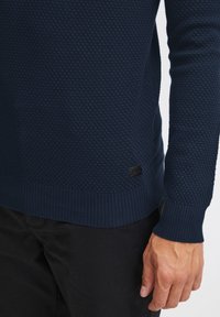 Navy blue textured sweater with a honeycomb pattern, ribbed cuffs and hem, featuring a small black logo tag at the lower side.