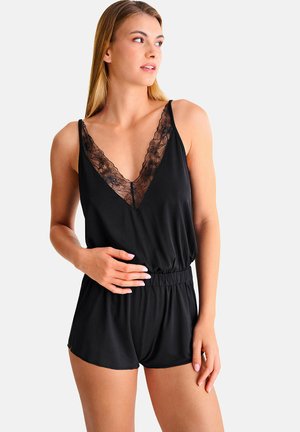 Femme portant une combinaison noire sans manches avec un décolleté en V orné de dentelle, debout avec une main sur la taille, regardant vers sa gauche sur un fond blanc.