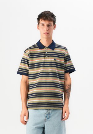 Young man wearing a striped short-sleeve polo shirt in navy, beige, white, and orange with light blue jeans against a plain white background.