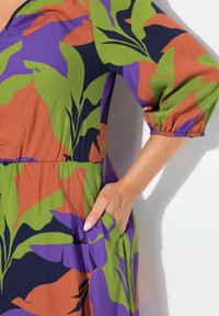 Bunt gemustertes Kleid mit einer Mischung aus grünen, lila und orangefarbenen Blattmustern. Aus leichtem Stoff gefertigt, mit tailliertem Schnitt und Taschen.