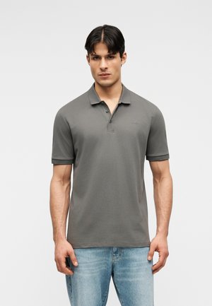 Fiatal férfi szürke rövid ujjú pólóingben és világoskék farmerben, egyszínű fehér háttér előtt áll.