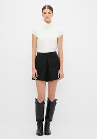 Top blanco ajustado con un cuello alto, combinado con una falda corta negra de pliegues que tiene bolsillos con solapa y botas de cuero negro hasta la rodilla.