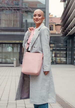 Růžová kabelka s hladkou texturou a nastavitelným popruhem, vystavená na světle šedém kostkovaném kabátu, v městském prostředí.