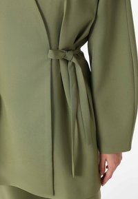 Olive green, soft fabric top with a tied waist detail, featuring long sleeves and a loose design, emphasizing comfort and versatility.