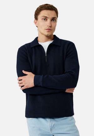 Junger Mann mit hellbraunen Haaren, trägt einen marineblauen, gerippten Zip-Up-Pullover über einem weißen Hemd und hellblauen Jeans, die Arme verschränkt.