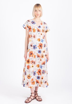 Žena v bielych midi šatách s oranžovými, modrými a žltými kvetinovými motívmi, krátkymi rukávmi a hnedými sandálmi na remienky na bielom pozadí.