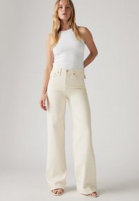 Calças de cintura alta e perna larga em ganga de cor creme com textura suave; apresentam bolsos frontais, fecho de botões e detalhes sutis de costura.