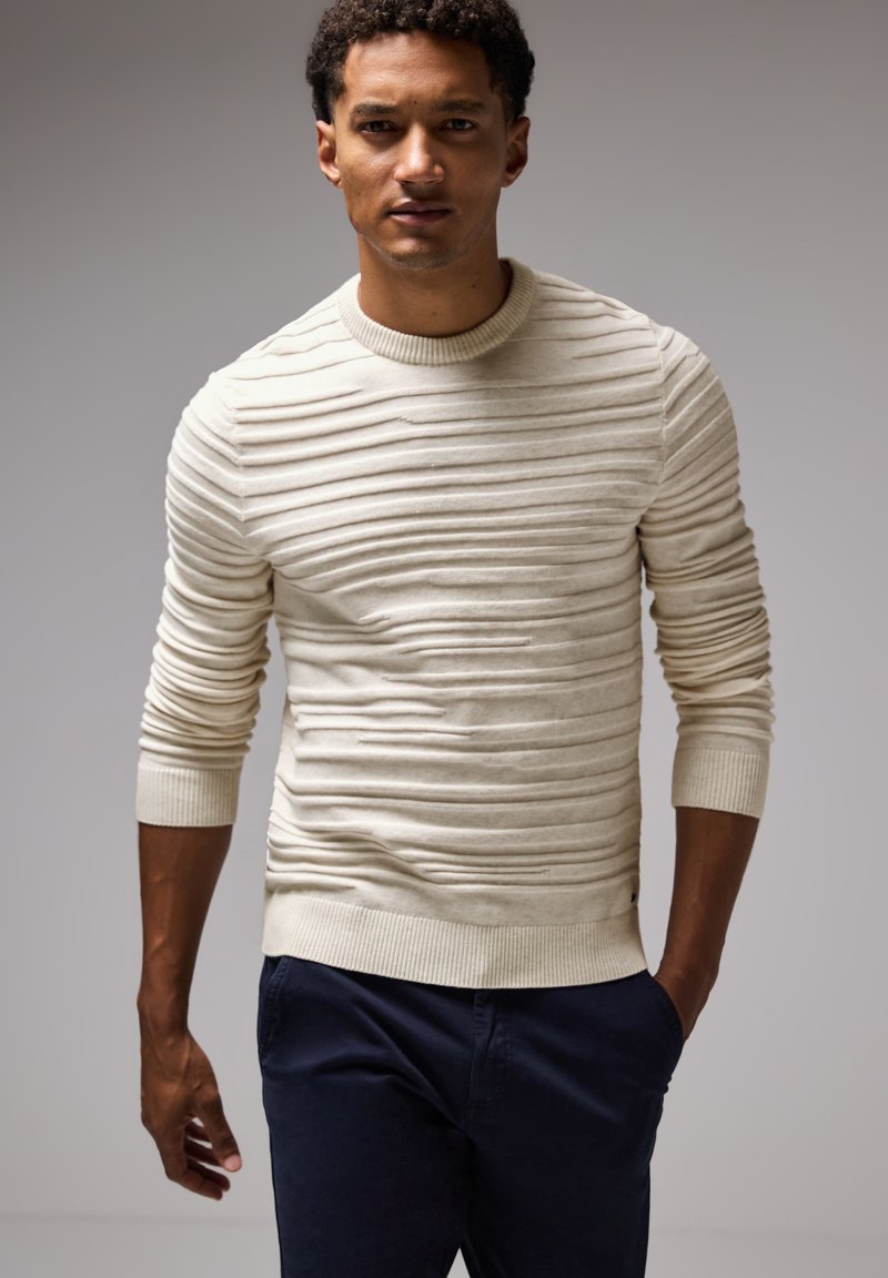 Stickad tröja i off-white, med texturerade horisontella ränder, ribbat rundhals och mudd. Kombinerad med marinblå byxor.