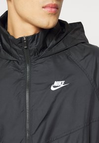 Melnā Nike jaka ar rāvējslēdzēju priekšpusē, kapuci un baltu ikonu kreisajā krūtī. Materiāls ir gluds, viegls un ūdensizturīgs.