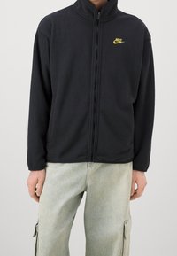 Svart fleecejacka med hög krage, hel dragkedja och fickor med dragkedja. Har en liten gul Nike-logotyp på vänster bröst.