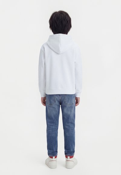 Παιδί που φοράει λευκή φούτερ με κουκούλα, μπλε τζιν και λευκά αθλητικά παπούτσια, στέκει με την πλάτη στραμμένη σε απλό λευκό φόντο.