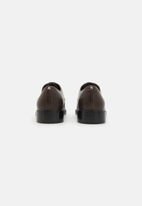 Brune lædersko med en blank finish, rund tå, lav sort hæl og sølvaccent nær bagsiden. Minimalistisk design.
