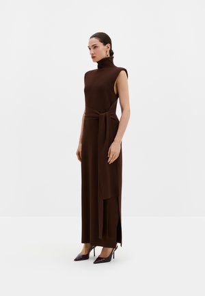 Femme portant une robe longue sans manches marron foncé à col montant avec des fentes latérales et une ceinture assortie à la taille, associée à des chaussures à talons aiguilles pointues.